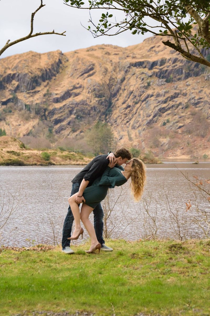 Man dipping his partner backward for a kiss beside a mountain lake