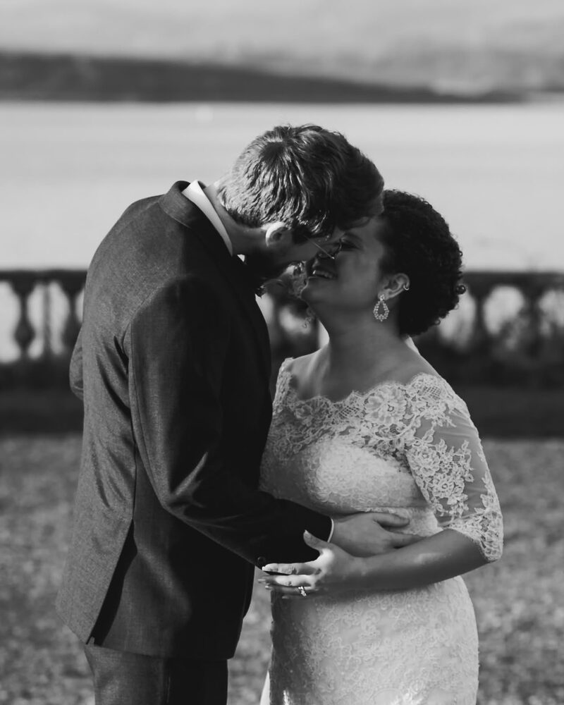 Black and white photograph of a bride and groom with foreheads touching beside the coast