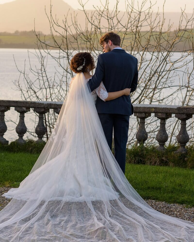 Bride and groom embracing on a terrace overlooking the water, her cathedral veil trailing behind